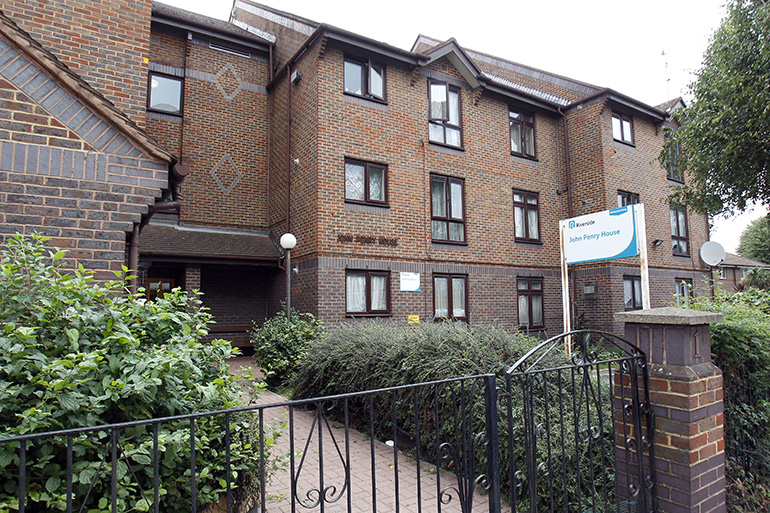 Exterior of a multi‑storey brick residential building with white‑framed windows, viewed through a black metal gate. A paved path leads to the main entrance, with low shrubs and planting along the frontage, and a sign reading “John Penry House” mounted near the doorway under an overcast sky.