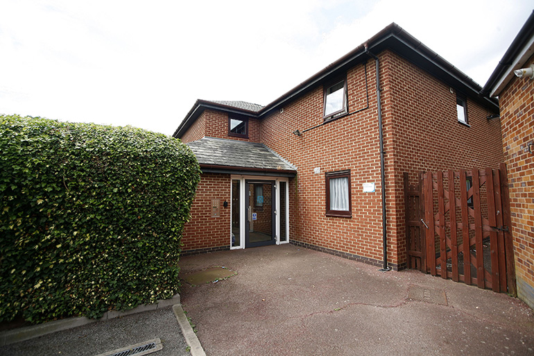 Exterior of a two‑storey red‑brick residential building with wooden‑framed windows and a pitched tiled roof, viewed from a paved entrance area. A glass entrance door sits beneath a small canopy, bordered by a tall hedge on one side and a wooden gate on the other, with the building extending to the right under a bright, overcast sky.