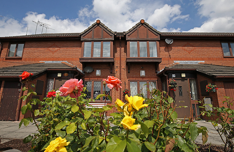Exterior of a row of red‑brick terraced houses with white‑framed windows and front doors. In the foreground, a flower bed of blooming pink and yellow roses sits beside a paved path, with hanging baskets by the entrances and a bright blue sky with scattered clouds above.