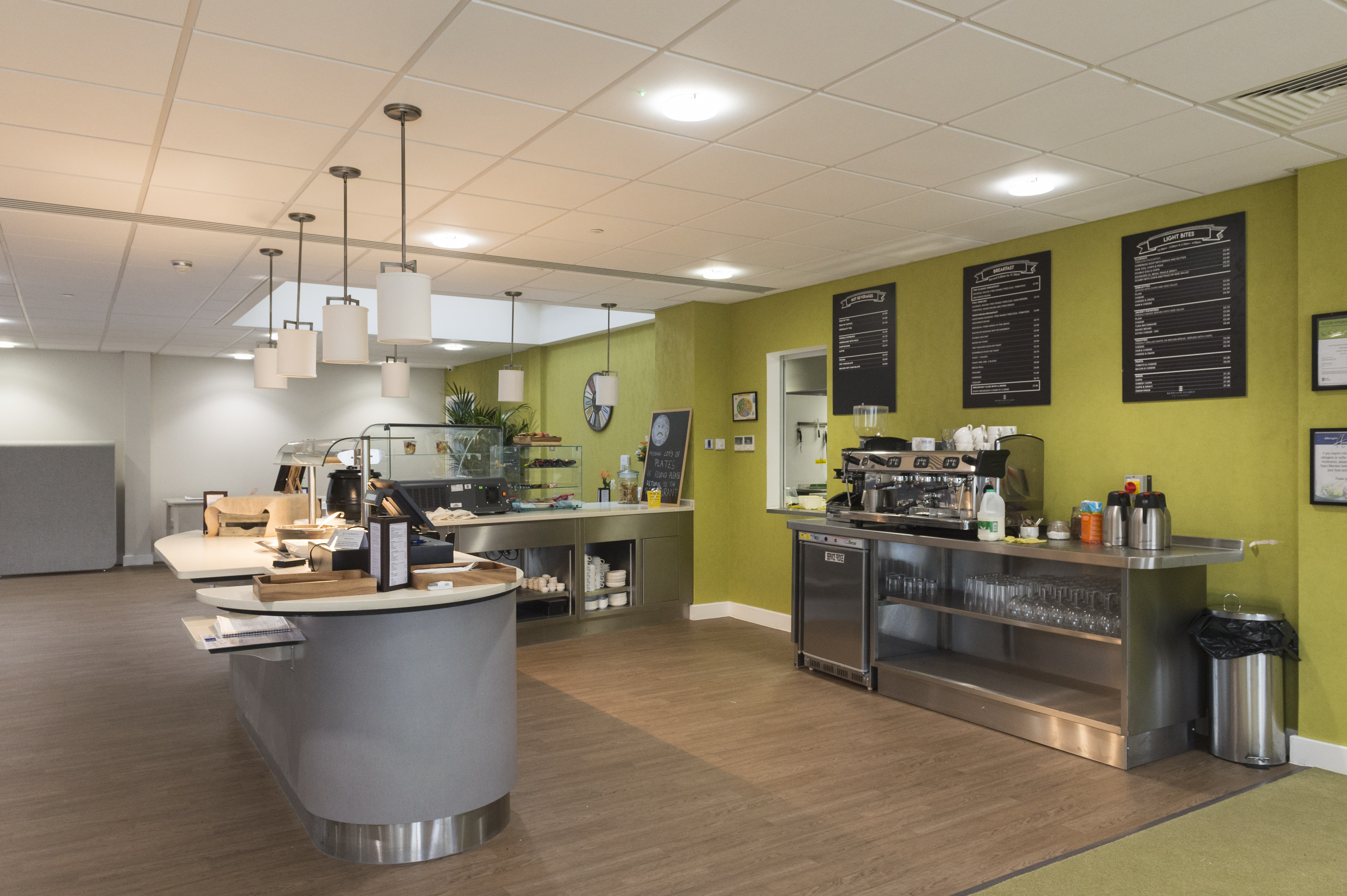 Café-style dining area with a serving counter, coffee machine, overhead menu boards, and display units, set against a bright green wall.