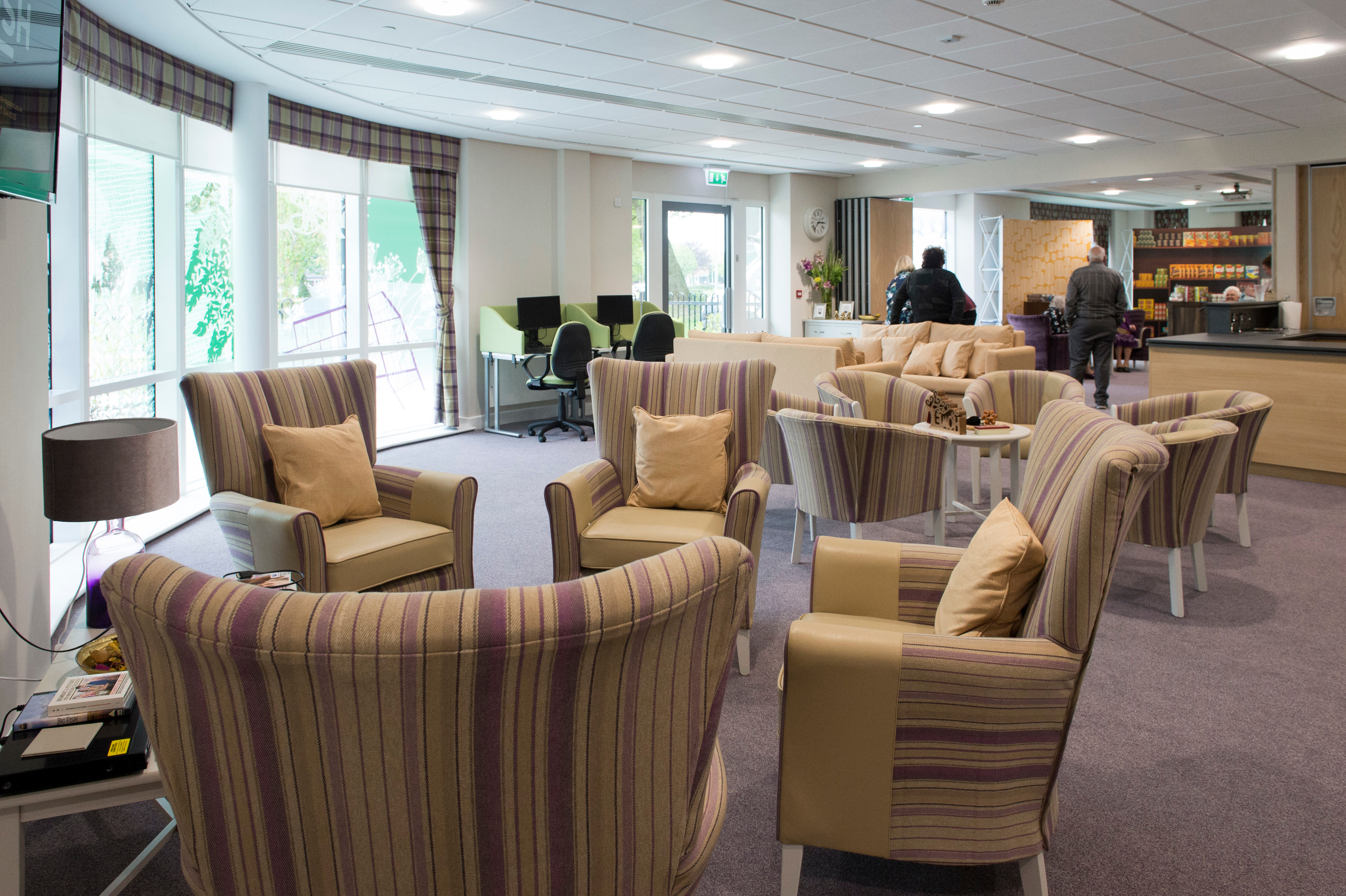 Communal lounge with armchairs and sofas, computer stations and a café area, with people using the shared space.