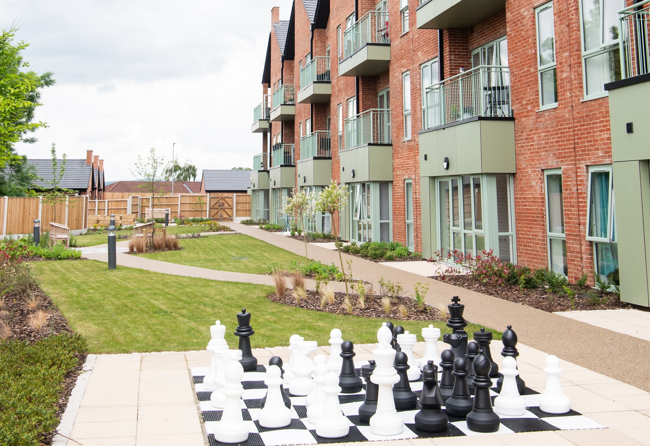 Newly built apartment buildings with balconies overlooking a landscaped shared garden. In the foreground, a large outdoor chess set is set out on a paved area, with lawns, paths and planting around the residential courtyard.