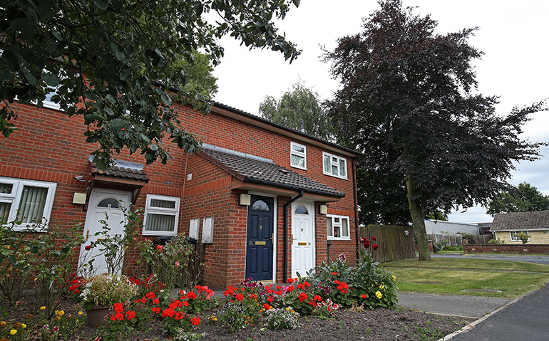 Exterior of a pair of red‑brick semi‑detached houses with small tiled porches and white‑framed front doors. A well‑kept flower bed with red and yellow plants lines the front of the properties, with a grassy verge, mature trees and neighbouring homes visible along the street.