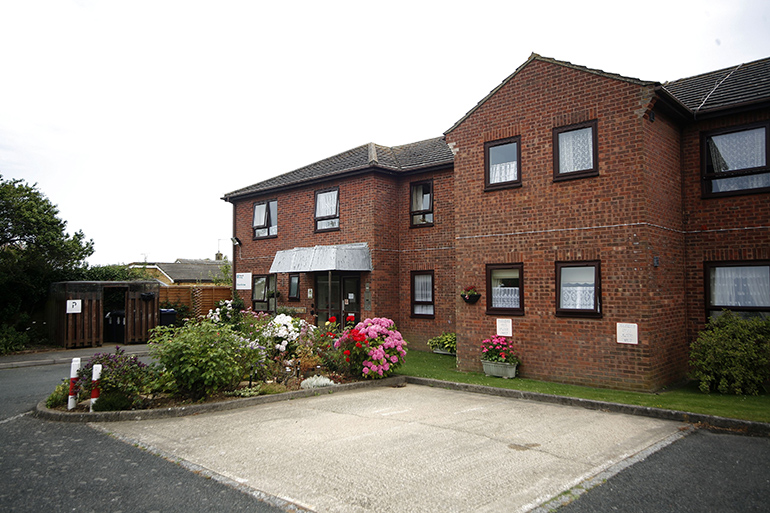 Exterior of a two‑storey red‑brick residential building with white‑framed windows and a central entrance canopy. A small landscaped garden with flowering shrubs sits beside a paved parking area in the foreground, with neighbouring buildings and fencing visible around the site under an overcast sky.