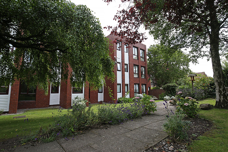Landscaped garden beside a red‑brick multi‑storey residential building with white‑clad sections. A paved pathway runs through flower beds and shrubs, with mature trees framing the scene and windows overlooking the shared green space.