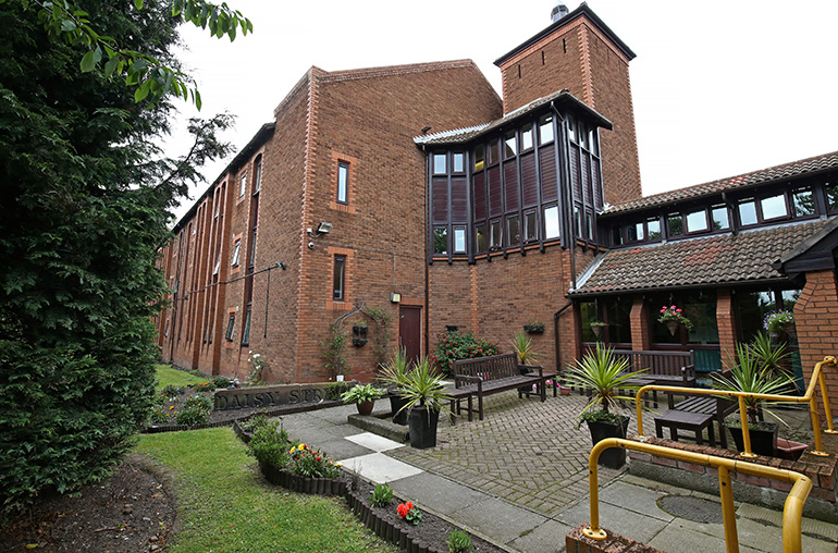 Exterior courtyard of a red‑brick residential building with a distinctive tower feature and enclosed glazed walkway. Paved seating areas with wooden benches and large planters sit among landscaped beds, with yellow handrails along steps and paths, and surrounding brick façades and windows overlooking the shared outdoor space.