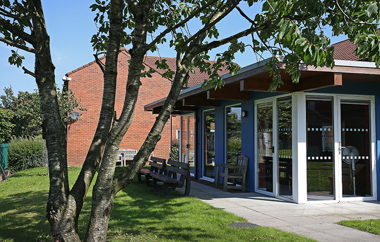 Communal garden area beside a single‑storey pavilion‑style building with large glass doors and blue exterior panels. Wooden benches sit on a paved patio, with a grassy lawn, mature trees and a red‑brick residential building visible in the background under clear daylight.