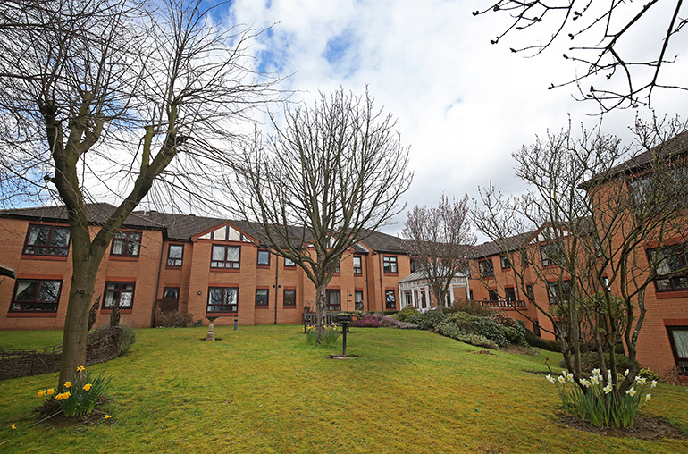 Wide communal garden courtyard surrounded by multi‑storey red‑brick residential buildings with white‑framed windows. A large central lawn is bordered by shrubs, small trees and spring flowers, with paved paths and a covered entrance visible in the background under a partly cloudy sky.