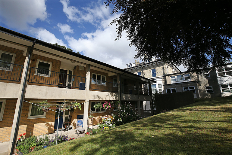 Exterior of a residential building on a sloping lawn, with cream‑coloured brick on the upper level and darker brick at ground level. A covered balcony walkway runs along the first floor, with flower displays and seating beneath. Mature trees frame the scene, with additional residential buildings visible in the background under a partly cloudy blue sky.