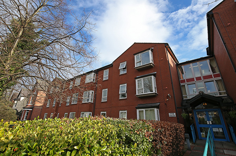 Exterior of a multi‑storey red‑brick residential building with white‑framed bay windows, partially screened by a neatly trimmed hedge in the foreground. A central entrance with blue double doors and handrails sits to the right, with mature trees and surrounding buildings visible under a partly cloudy blue sky.