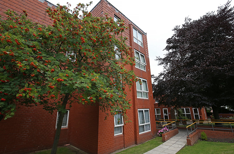 Exterior of a multi‑storey red‑brick residential building with white‑framed windows, set within landscaped grounds. A paved path and access ramp with yellow handrails lead to the entrance, with mature trees, flowering shrubs and a grassy lawn around the building, under an overcast sky.