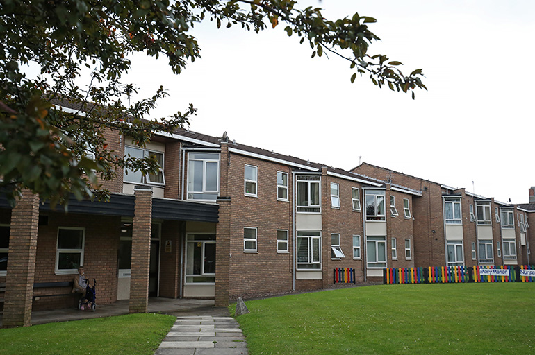Exterior of a two‑storey brick residential building with white‑framed windows, viewed across a wide communal lawn. A paved path leads to a covered entrance on the left, with a bench nearby. A colourful fence on the right, and trees and planting frame the shared outdoor space under an overcast sky.