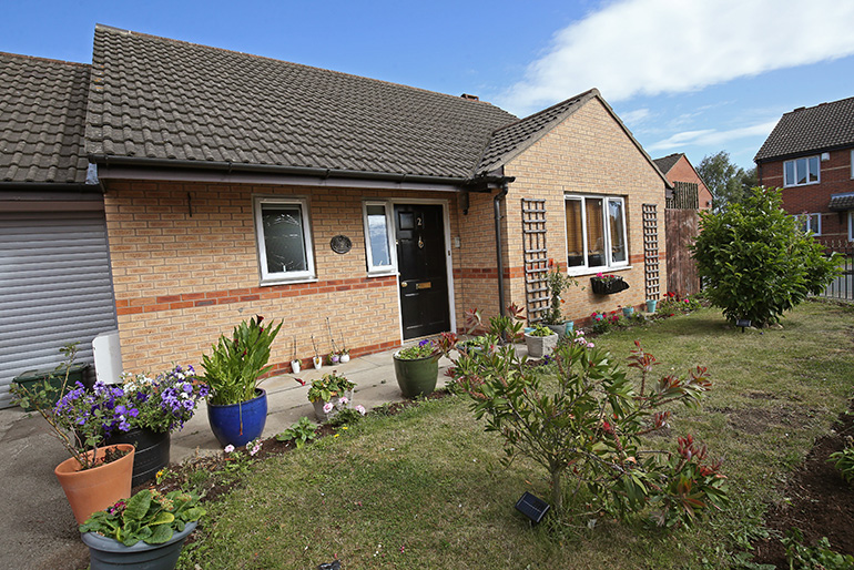 Exterior of a single‑storey light‑brick bungalow with white‑framed windows and a dark tiled roof. The front garden is neatly landscaped with potted plants, flower beds and small shrubs, with a paved path leading to a black front door. Trellis panels and neighbouring homes are visible to the side under a bright blue sky.