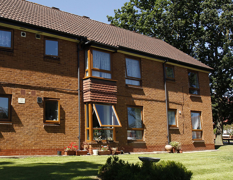 Exterior of a two‑storey red‑brick residential building with wooden‑framed windows and a pitched tiled roof. The building faces a neatly maintained lawn, with potted plants and small shrubs along the frontage and a mature tree casting shade across the grass under bright sunlight.