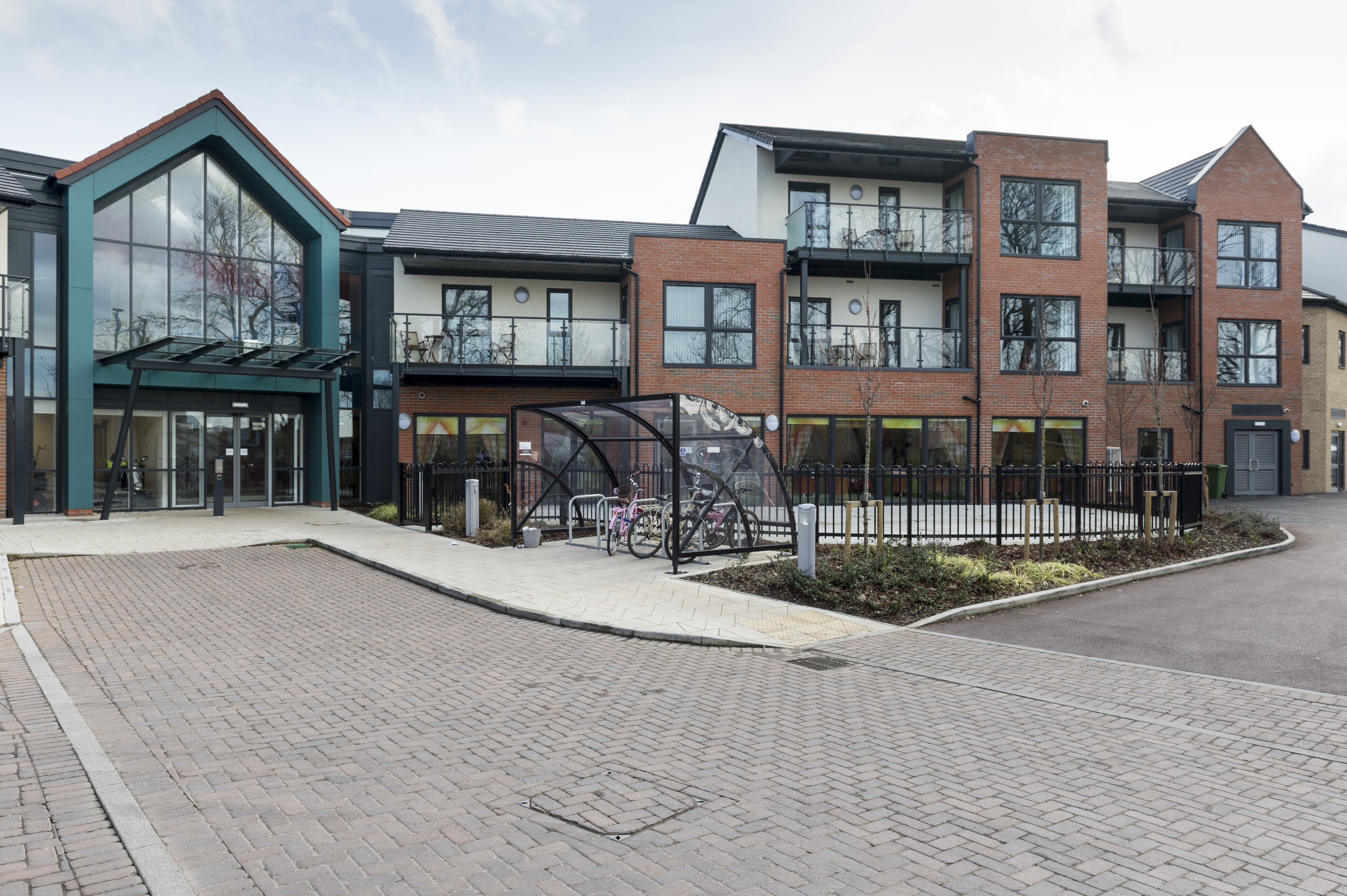 Exterior view of a modern residential building with brick and glass façades, balconies, landscaped paths, and a covered bicycle storage area at the entrance.