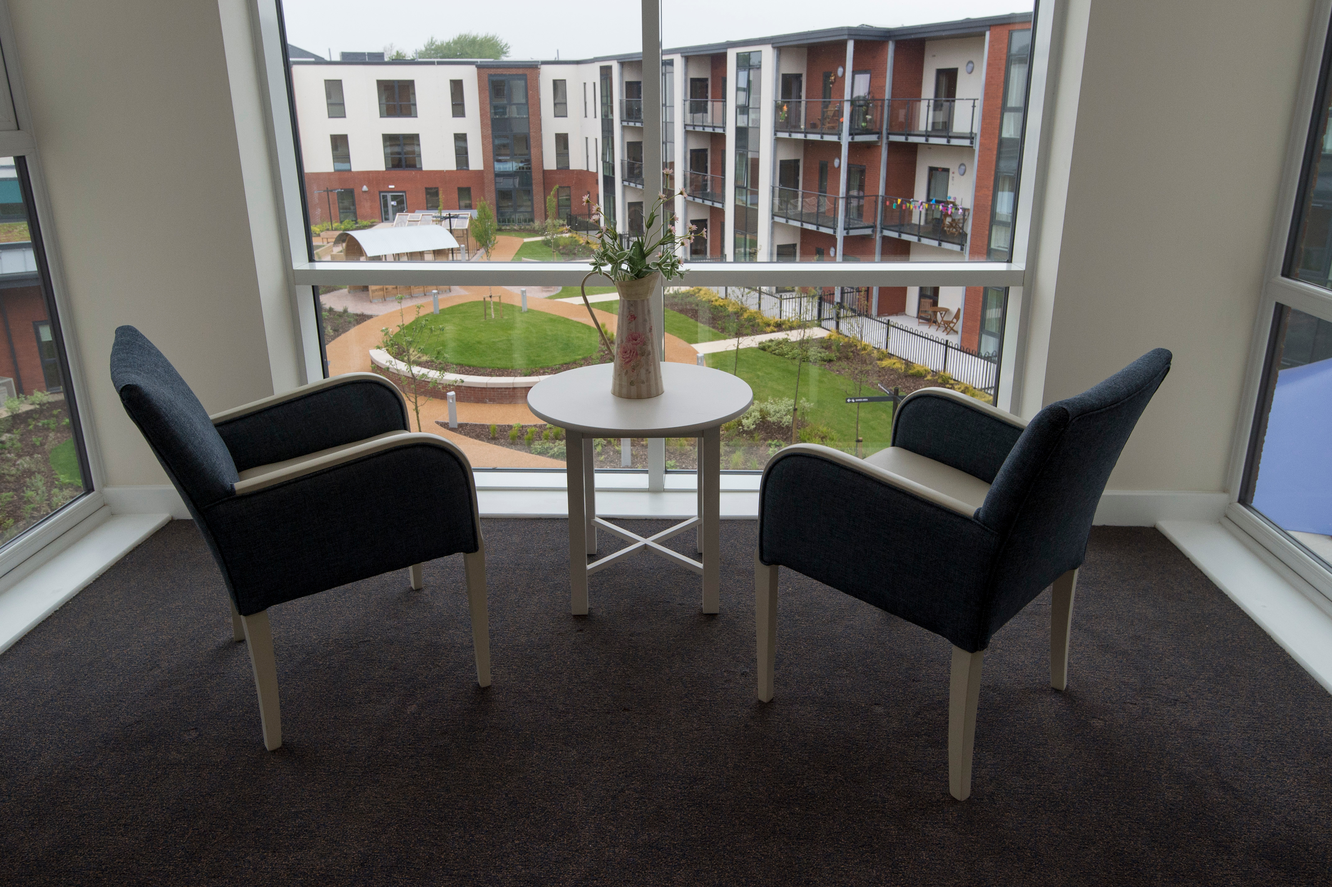 Small seating area with two armchairs and a table positioned by a large window overlooking landscaped gardens and apartments.