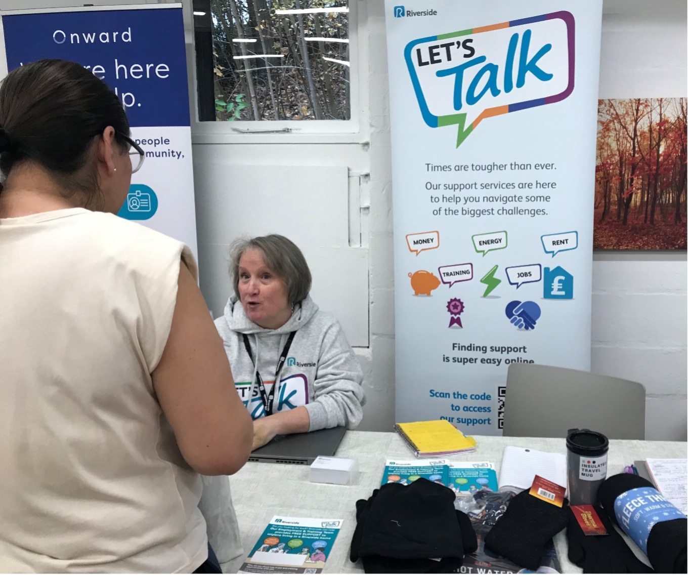 Let's Talk Murdishaw Community Event A person speaks with a staff member seated behind an information table at a community event. A Riverside “Let’s Talk” banner stands behind the table, promoting support with money, energy, rent, training and jobs. Leaflets and branded items are displayed on the table.