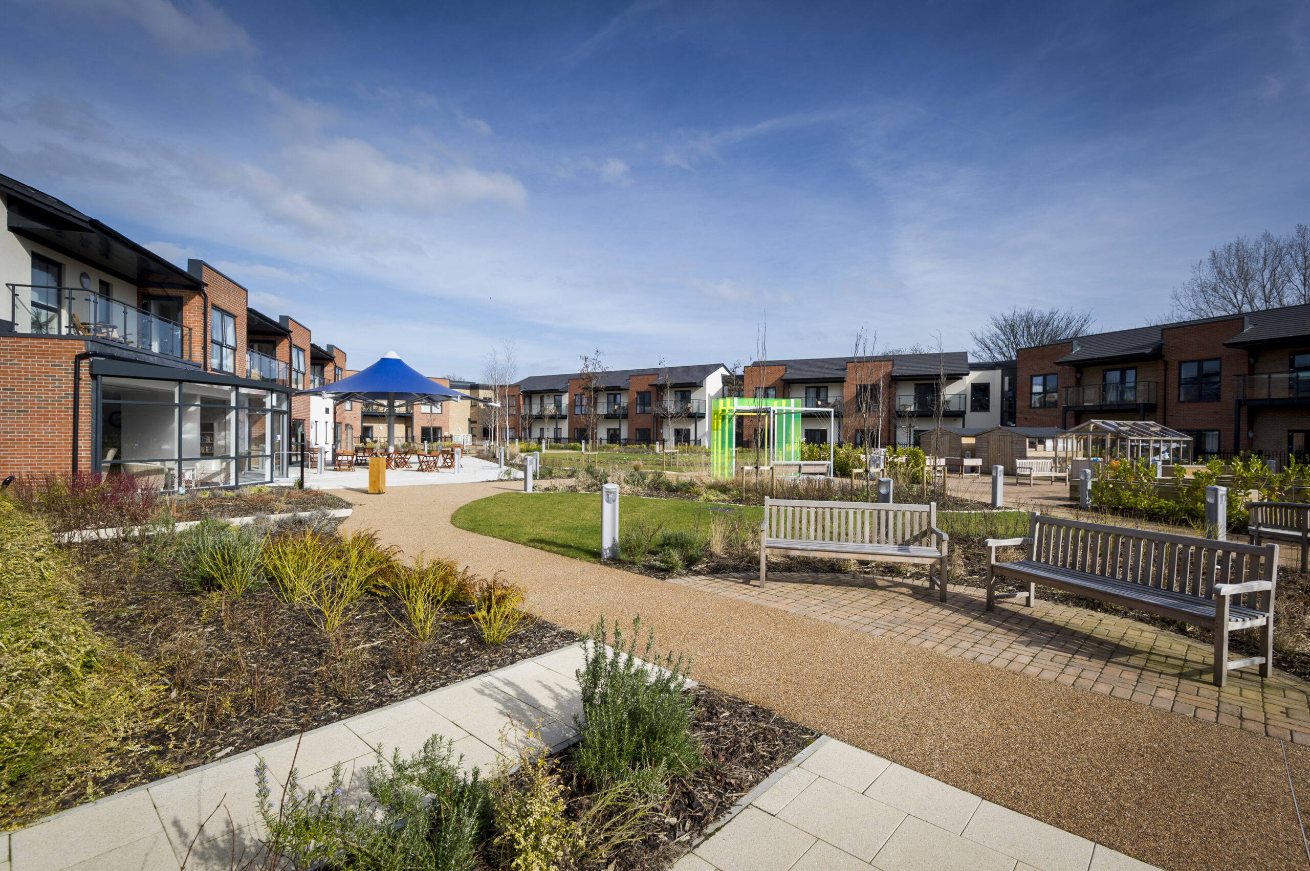 Landscaped residential courtyard with pathways, benches and planted gardens, surrounded by modern low‑rise apartment buildings. A small play area and shared outdoor seating are visible under a clear blue sky.