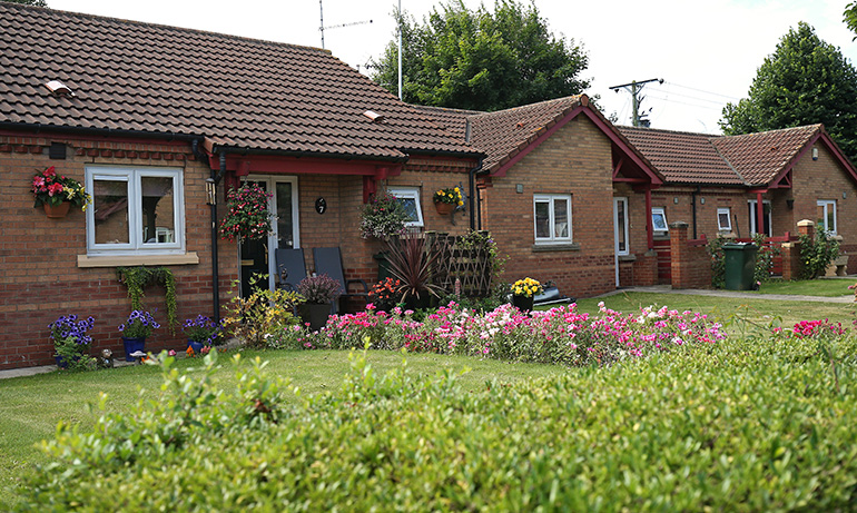 Row of single‑storey red‑brick bungalows with pitched tiled roofs and white‑framed windows. Colourful hanging baskets and flower beds line the front gardens, with a neatly trimmed hedge in the foreground and trees visible behind the homes.