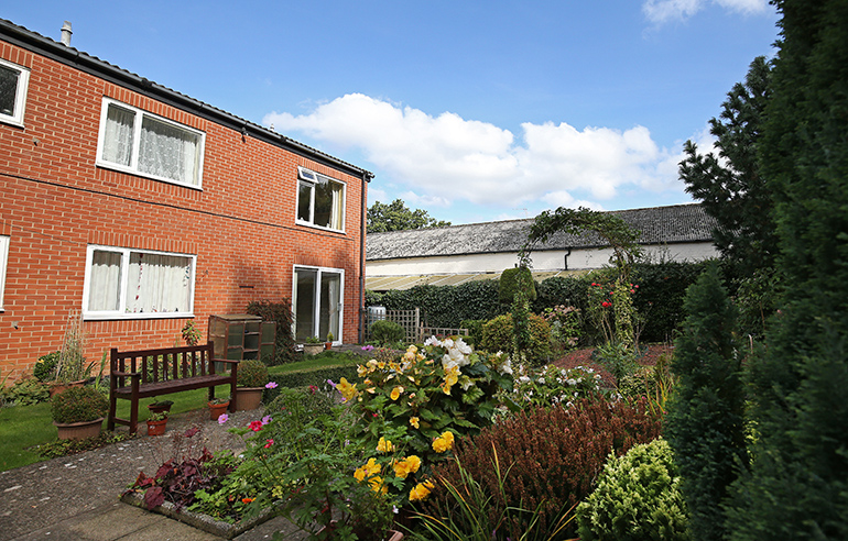 Landscaped communal garden beside a two‑storey red‑brick residential building with white‑framed windows. A wooden bench sits near a paved path, surrounded by well‑maintained flower beds and shrubs in bloom, with a neatly kept lawn and hedging under a blue sky with scattered clouds.