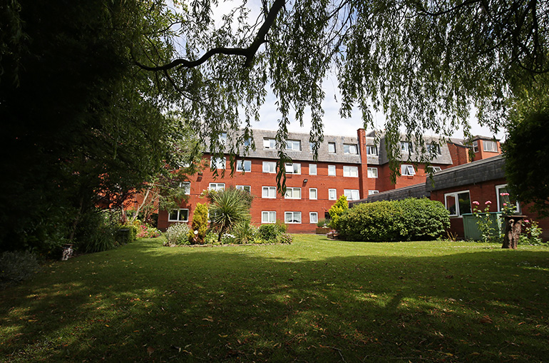 Wide communal garden in front of a multi‑storey red‑brick residential building, with a large grassy lawn, mature trees and shrubs creating shaded areas. The building has white‑framed windows and upper‑floor dormer windows, with landscaped planting and seating areas around the shared green space.