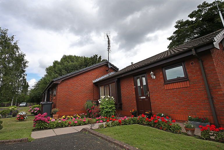 Exterior of a single‑storey red‑brick bungalow with white‑framed windows and a dark front door, set within a well‑kept garden. A paved path leads to the entrance, bordered by neatly arranged flower beds with colourful plants and shrubs, a trimmed lawn, and mature trees nearby under a cloudy sky.