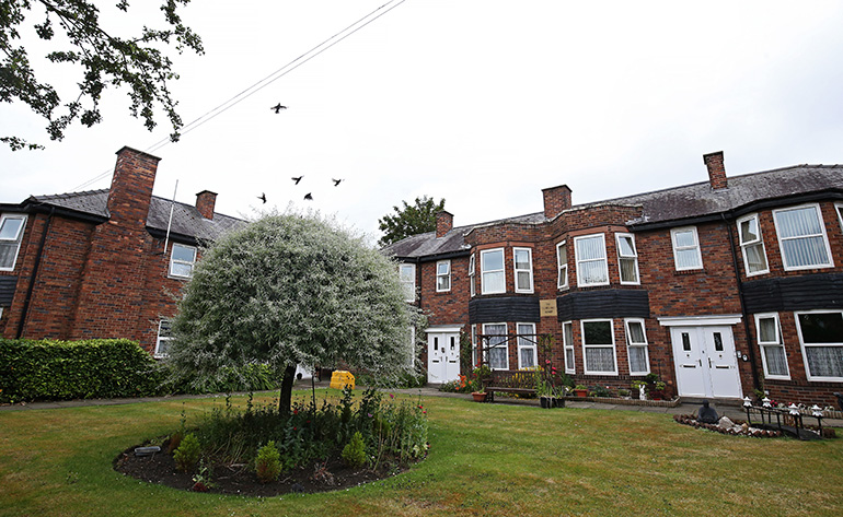 Communal garden in front of a row of red‑brick residential buildings with white‑framed bay windows and front doors. A circular planted flower bed with a small tree sits at the centre of a neatly mown lawn, with benches, shrubs and a paved path, and birds flying above the rooftops under an overcast sky.