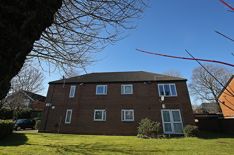 Exterior of a two‑storey red‑brick residential building with white‑framed windows, viewed across a neatly maintained lawn. Shrubs and small bushes sit near the base of the building, with leafless tree branches framing the scene against a clear blue sky.