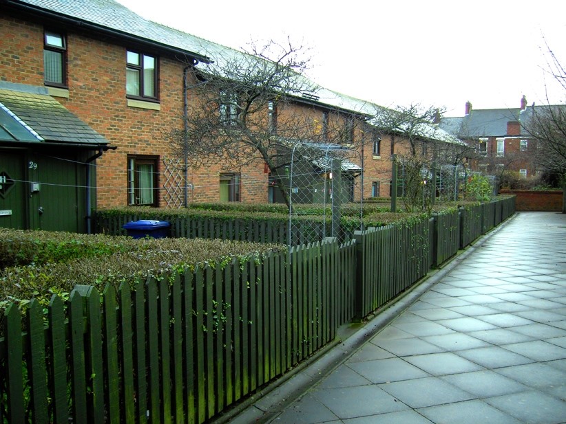 Row of red‑brick residential houses with white‑framed windows, set behind low hedges and a dark wooden picket fence. A wide paved footpath runs along the front of the homes, with small front gardens, trellis panels and leafless trees visible under an overcast sky.