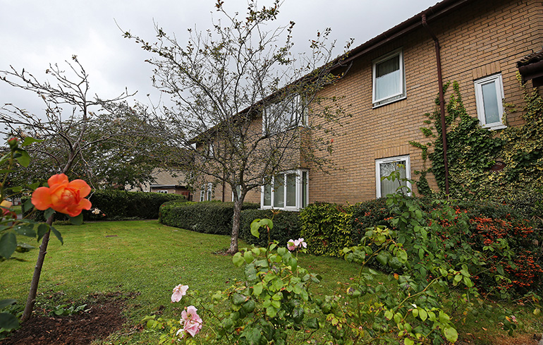 Exterior of a two‑storey residential building with light brown brick walls and white‑framed windows, viewed across a landscaped garden. A neatly maintained lawn, shrubs and rose bushes fill the foreground, with a small tree and climbing plants along the building, under an overcast sky.