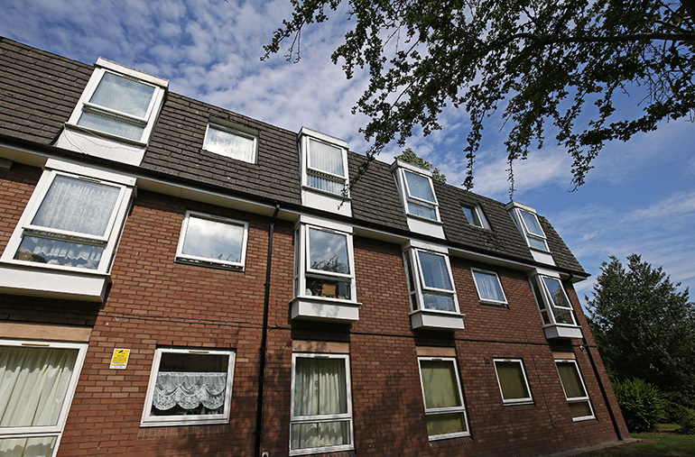 Exterior of a multi‑storey red‑brick residential building with white‑framed windows and dormer windows set into a dark tiled roof. The building is viewed from below, with leafy tree branches framing the top of the image and a blue sky with scattered clouds in the background.