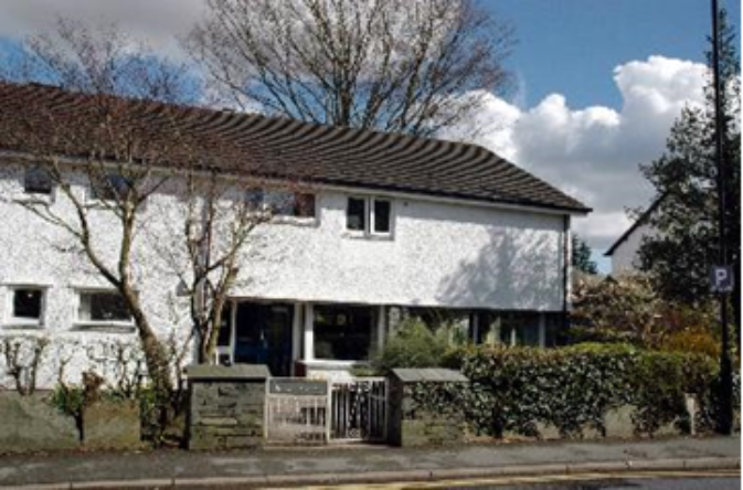 Exterior of a two‑storey white‑rendered residential building with white‑framed windows and a pitched tiled roof, set back from the street behind a low stone wall and hedge. A small gated entrance leads to the front area, with mature trees nearby and a blue sky with scattered clouds overhead.