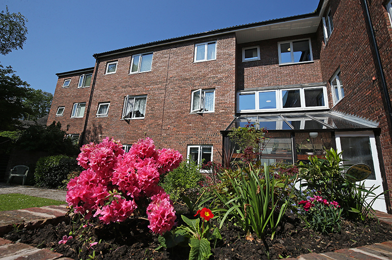 Exterior of a multi‑storey red‑brick residential building with white‑framed windows, viewed from a landscaped communal garden. Pink flowering plants and green foliage fill the foreground, with a paved path, lawn and a glass‑roofed conservatory visible beside the building under bright daylight.