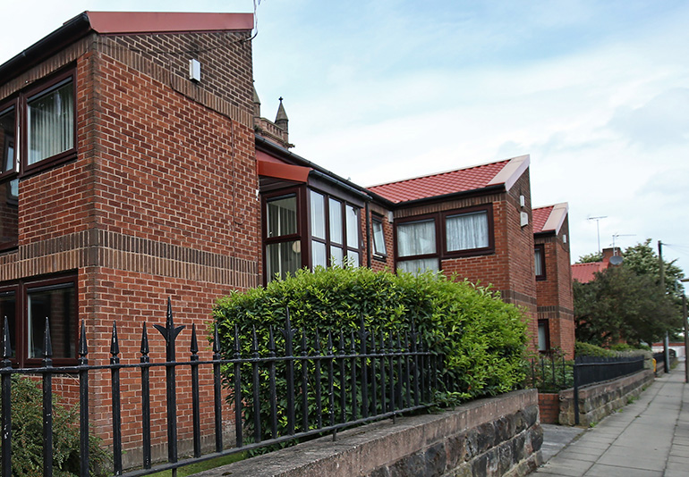 Exterior of a red‑brick residential building set along a street, with wooden‑framed windows and pitched tiled roofs. A black metal fence and low stone wall run along the pavement in the foreground, with hedges and shrubs beside the building and neighbouring homes visible under a bright, lightly clouded sky.
