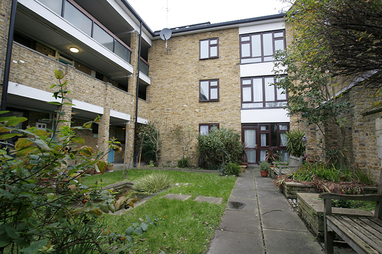 Communal courtyard beside a multi‑storey light‑brick residential building with wooden‑framed windows and balconies. A paved path runs through a small lawn with shrubs and planted beds, leading past wooden benches and seating areas, with mature trees and greenery around the shared outdoor space under overcast daylight.