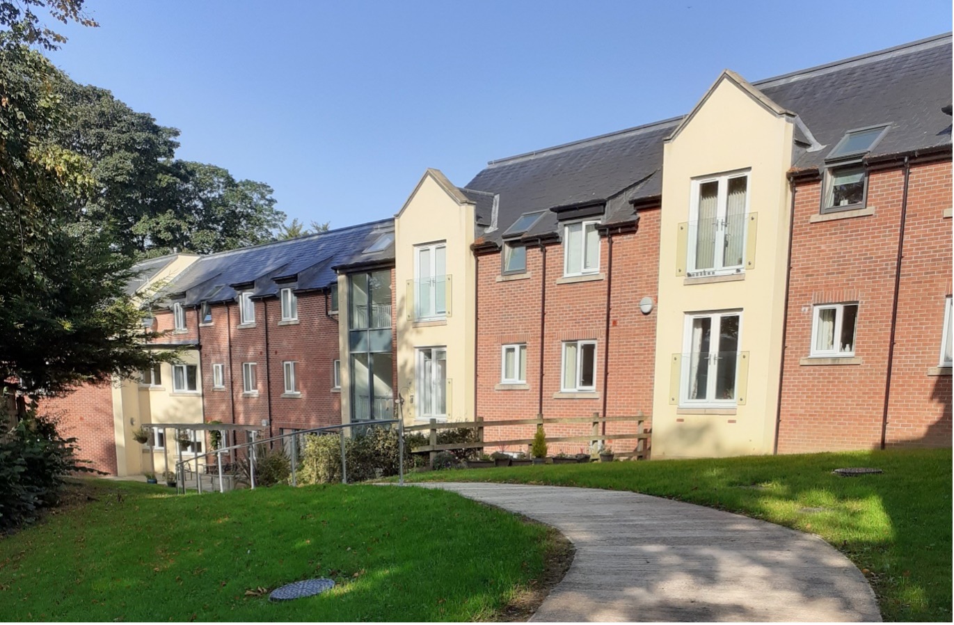 Exterior of a two‑storey red‑brick residential building with cream‑coloured rendered sections and white‑framed windows. A curved paved pathway leads through a neatly maintained lawn, with a ramp and handrail providing step‑free access, and mature trees and shrubs surrounding the development under a clear blue sky.