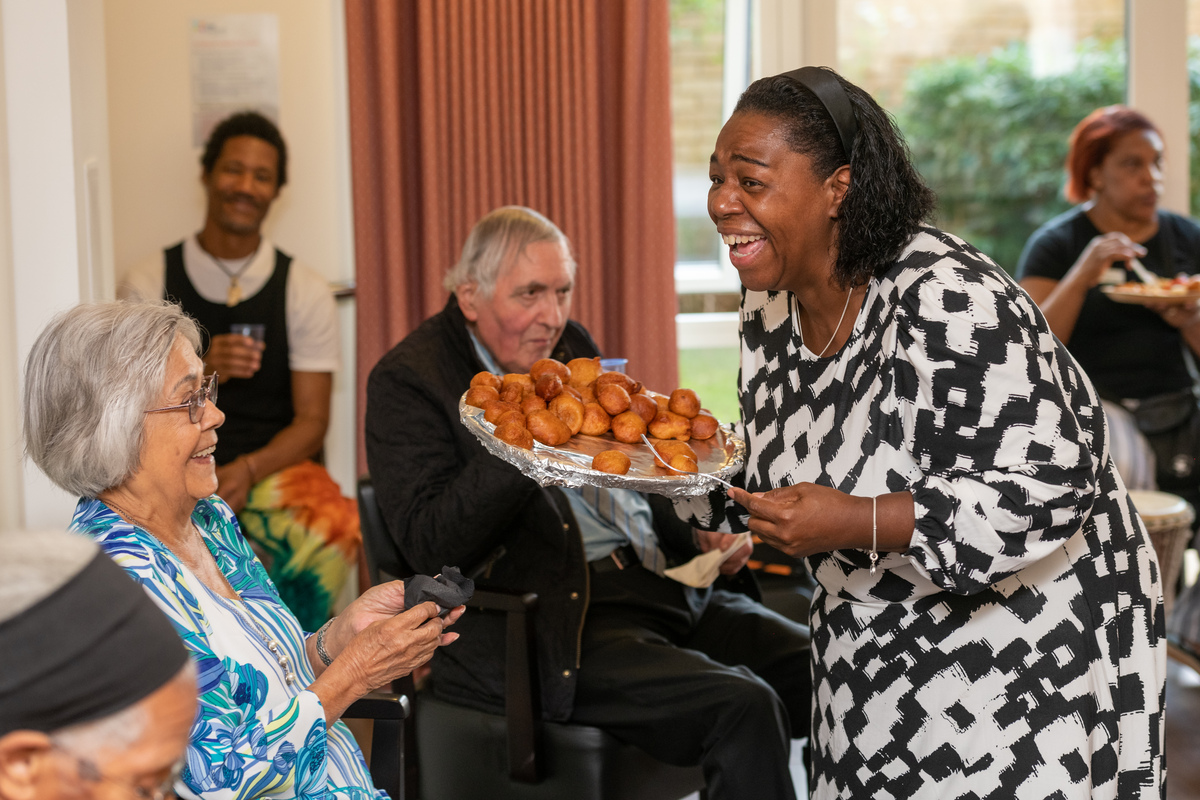 Person holding a tray of small round snacks, offering them to a seated group of adults in a communal indoor setting, with others sitting and eating in the background.