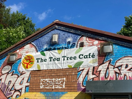 Tea Tree Community Cafe Exterior of The Tea Tree Café building with a colourful graffiti-style mural. A sign on the front reads “The Tea Tree Café – A Murdishaw Community Project,” with trees and greenery visible around the building under a blue sky.