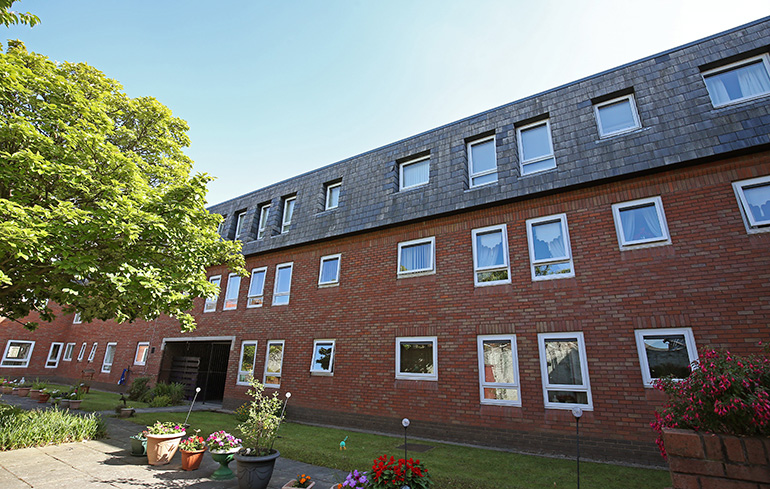 Exterior of a multi‑storey red‑brick residential building with rows of white‑framed windows. The building overlooks a landscaped communal garden with lawns, potted plants and flower beds, with a tree to one side and a clear blue sky above.