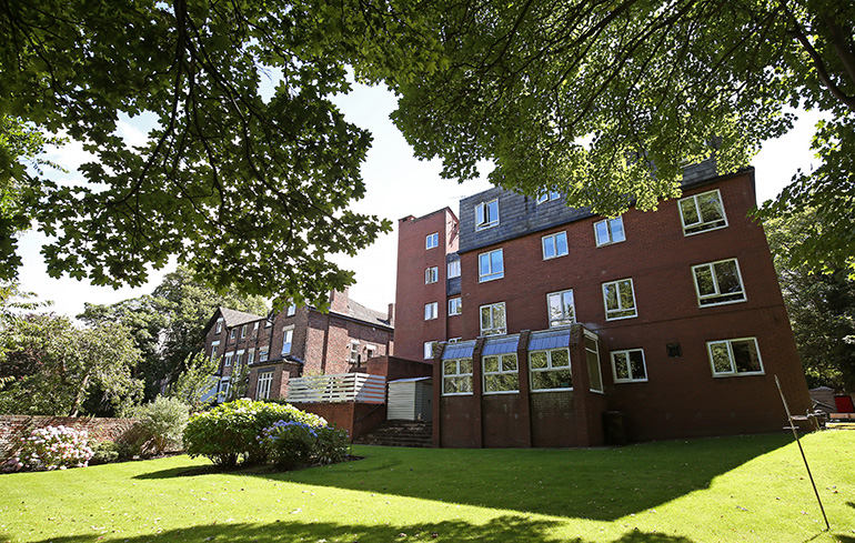 Red‑brick multi‑storey residential building set within landscaped grounds, viewed across a well‑kept lawn. Mature trees create dappled shade in the foreground, with shrubs and flower beds near the building and windows overlooking the shared green space.