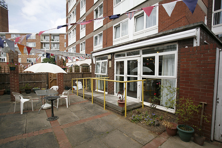 Paved outdoor seating area beside a red‑brick residential building, with white‑framed doors and windows opening onto the space. Tables and chairs sit under a large parasol, with bunting strung overhead, potted plants along the edges, and yellow handrails leading to the entrance.