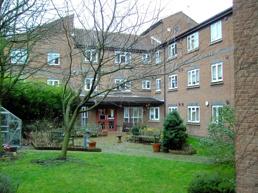 Communal garden courtyard outside a multi‑storey red‑brick residential building with white‑framed windows and red entrance doors. A paved seating area with wooden benches sits among lawns, shrubs and small trees, with planting beds and garden features arranged around the shared outdoor space.