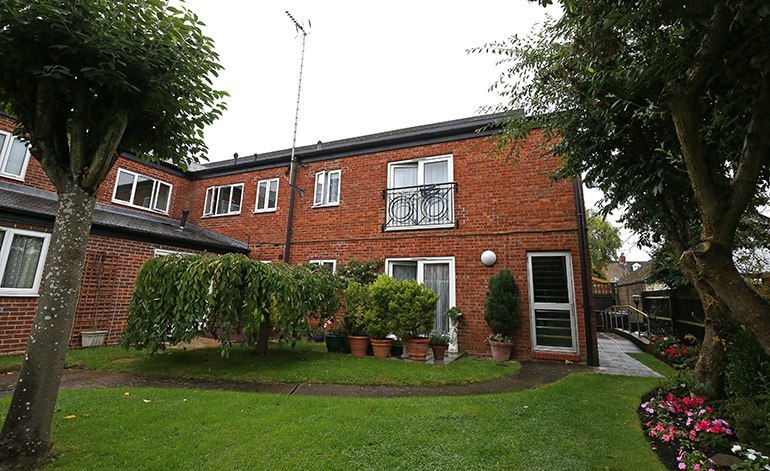 Communal garden outside a two‑storey red‑brick residential building with white‑framed windows and a small upper‑floor balcony. A curved paved path runs through a neatly kept lawn, with potted plants, shrubs and flowering beds near the building, and trees framing the shared outdoor space.