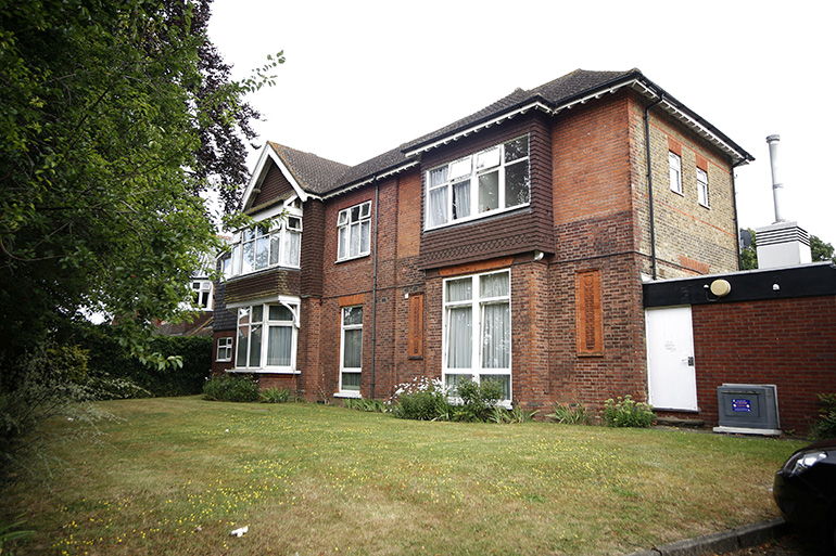 Exterior of a large two‑storey red‑brick residential building with white‑framed windows and decorative brick detailing. The building is set behind a gently sloping grass lawn with trees and shrubs to the side, and a small single‑storey extension visible on one end, under an overcast sky.