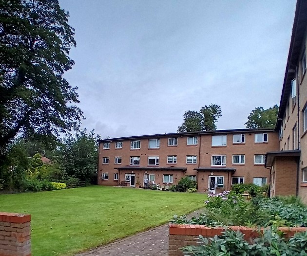 Communal garden behind a two‑storey red‑brick residential building with white‑framed windows. A wide grassy lawn stretches along the front of the homes, with a paved path, shrubs and planted borders, and mature trees surrounding the shared outdoor space under a cloudy sky.