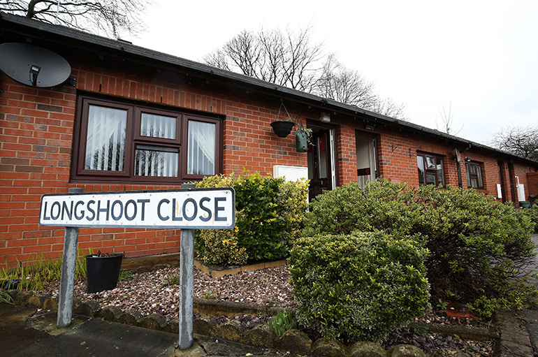 Exterior of a single‑storey red‑brick residential building with white‑framed windows, viewed from a landscaped front garden. A street sign reading “Longshoot Close” stands in the foreground beside low shrubs and gravel beds, with hanging baskets by the entrances and leafless trees behind the building under an overcast sky.