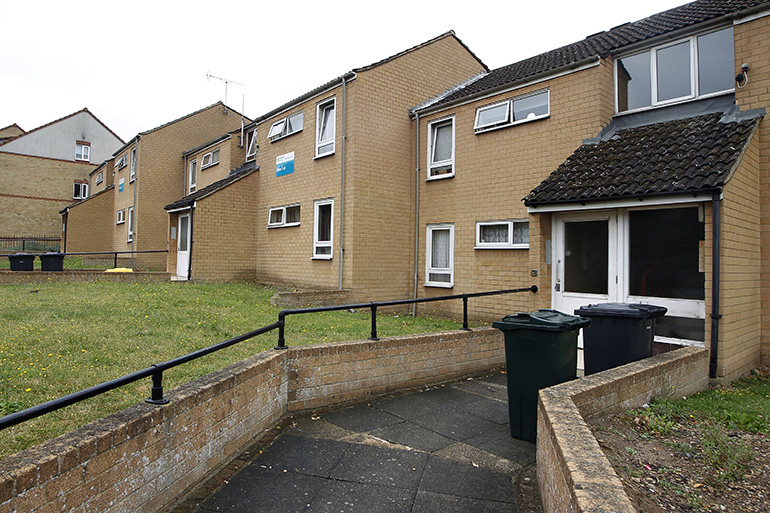 Exterior of a two‑storey light‑brick residential building with white‑framed windows, viewed from a paved access path with low brick walls on either side. Wheelie bins are positioned near a recessed entrance on the right, with a metal handrail running along the pathway, a grassed area to the left, and additional residential blocks visible under an overcast sky.