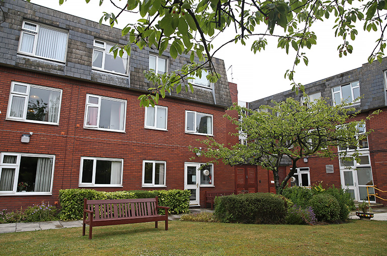 Communal garden courtyard outside a multi‑storey red‑brick residential building with white‑framed windows. A wooden bench sits on a neatly maintained lawn in the foreground, with shrubs, small trees and paved paths around the garden, and entrances and windows overlooking the shared outdoor space under an overcast sky.