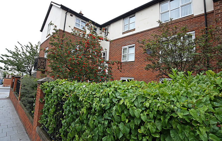 Corner view of a multi‑storey red‑brick residential building with white‑rendered upper sections and white‑framed windows. A black metal balcony projects from the first floor, and a street sign on the brick wall reads “Brownlow Hill.” A black metal fence and tree branches frame the foreground, with the building rising against a cloudy sky.