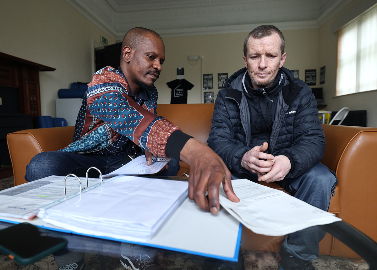 Two men sitting on a sofa reviewing documents together, one pointing to paperwork in an open binder on a coffee table while the other looks on thoughtfully, suggesting a discussion or support meeting.